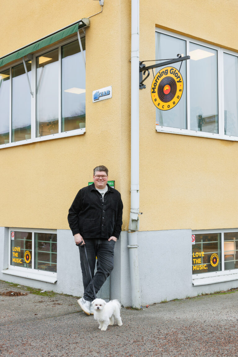 Ägare utanför Morning Glory Records i Katrineholm med liten vit hund, vinylbutik i gul hörnfastighet.